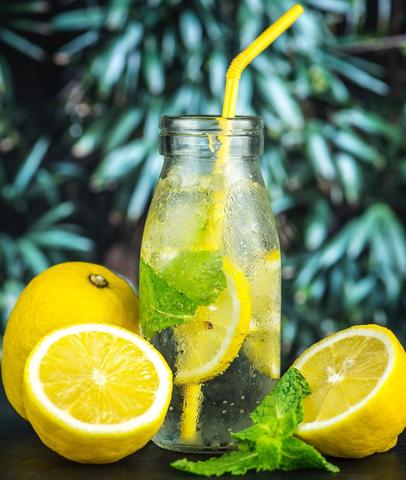Water with wedges of lemon and a green leaf in a clear glass bottle with yellow straw set on table. Surrounded with one lemon cut in half with a green leaf between them and one whole lemon and palm tree leaves as a backdrop. 