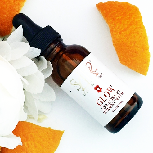 SyllyS of Switzerland GLOW concentrated vitamin c serum Face Serum laying down on a white background surrounded by orange peels along with an off white fluffly rose that is gently touching it. It is in a clear brown glass bottle with a black dropper.