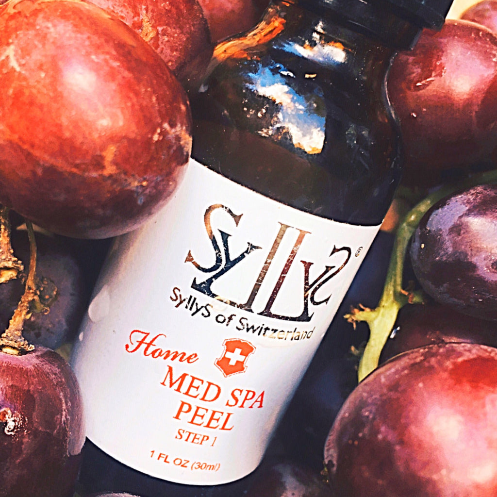 A closeup of "Home Med Spa Peel Step 1” brown glass bottle with a black cap laying down in a bed of red lush grapes. The logo Syllys of Switzerland is printed in gold on a white label, below it the title "Home Med Spa Peel" is printed in red and below it 