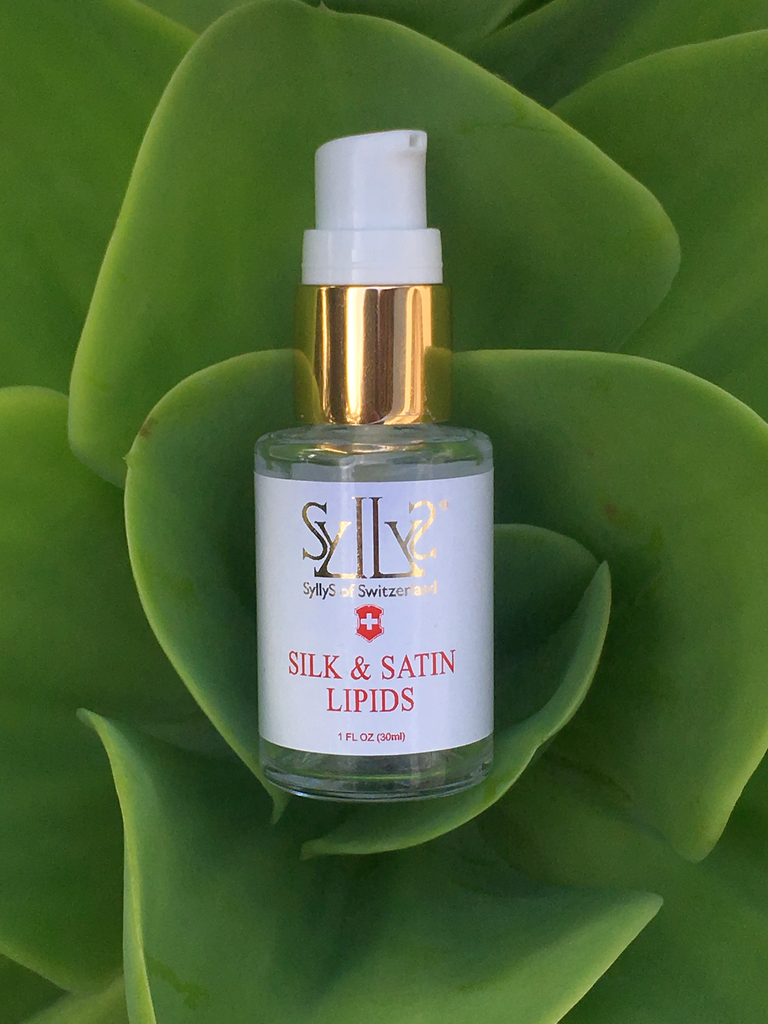 SyllyS Silk & Satin Lipids in a glass clear pump bottle with gold rim with white label with gold logo and title product printed in red below. It is laying down in the heart of a silky looking green plant. 